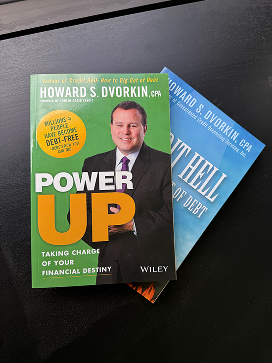 Two personal finance books by Howard S. Dvorkin, titled 'Power Up' and 'Credit Hell,' displayed on a black surface.