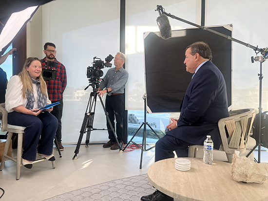 Howard Dvorkin being interviewed on camera in a professional video shoot setup with crew and lighting equipment, seated in front of a scenic window backdrop.