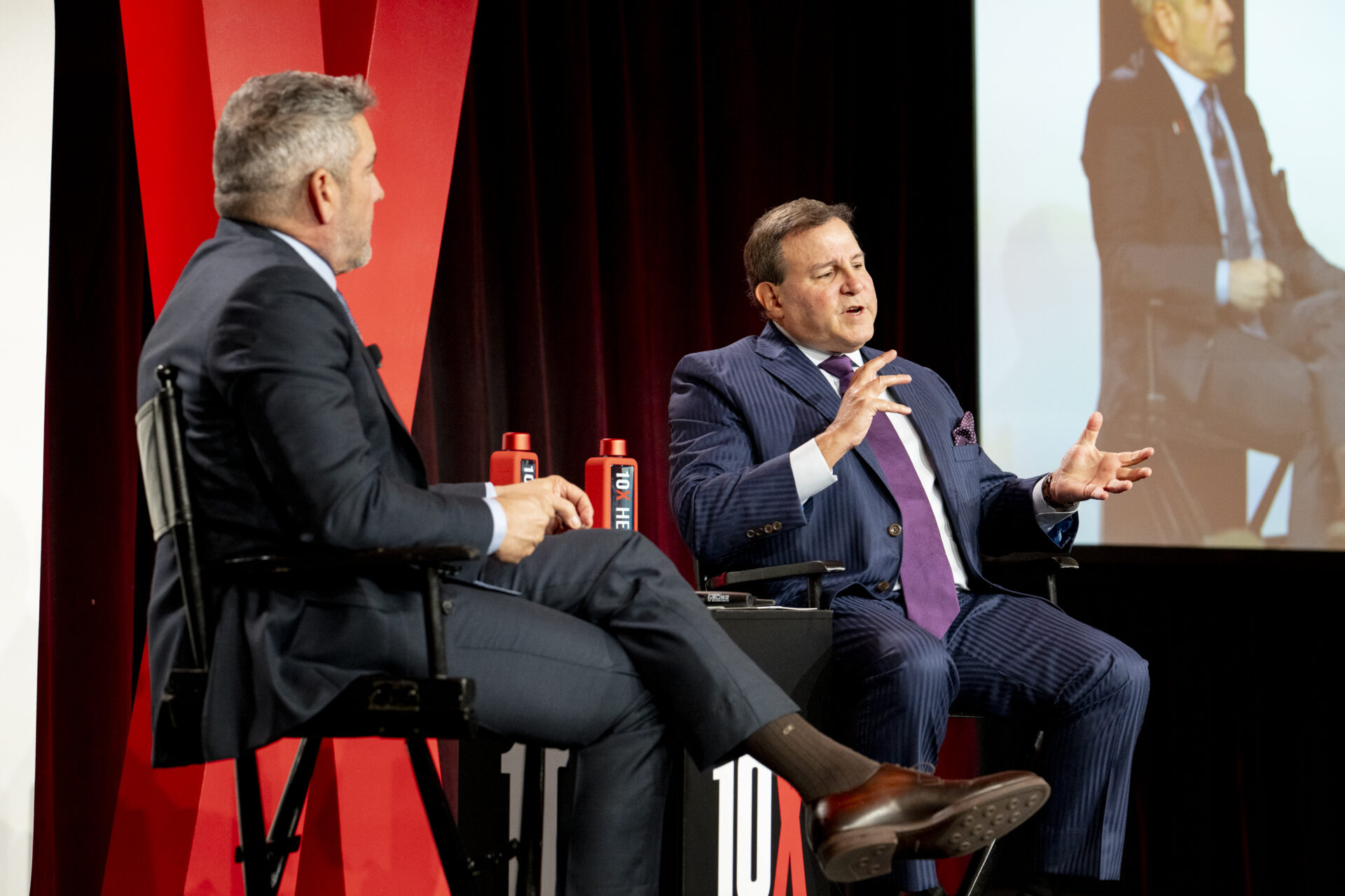 Howard Dvorkin speaking on stage during a live panel discussion, seated beside a moderator, with a 10X Health backdrop and screen projection in the background.