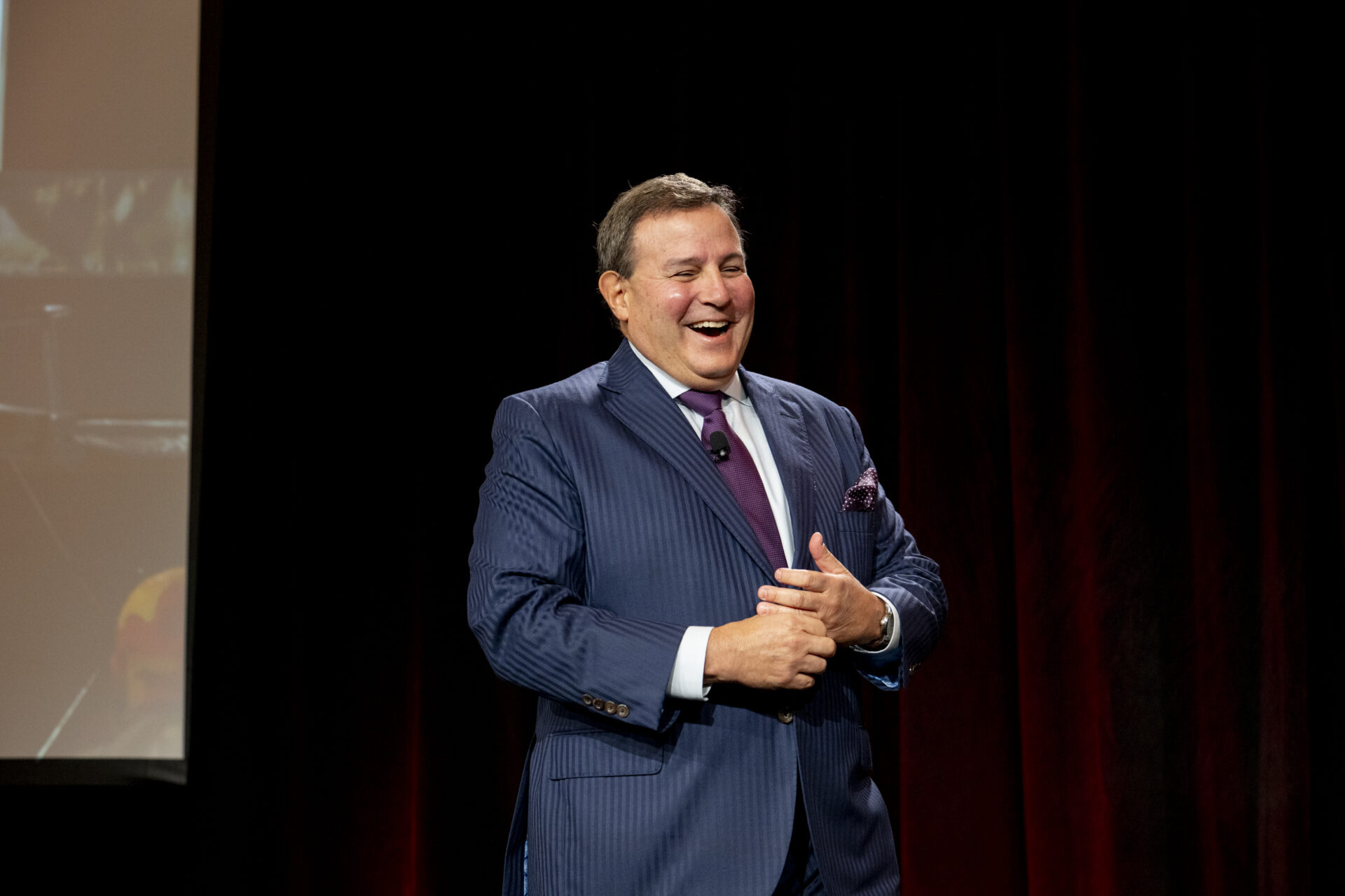 Howard Dvorkin, financial expert and industry leader, speaking confidently on stage during a keynote event, wearing a pinstripe suit and purple tie against a dark backdrop.